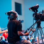 Two cameramen operate professional video cameras at an indoor event with a lively audience.